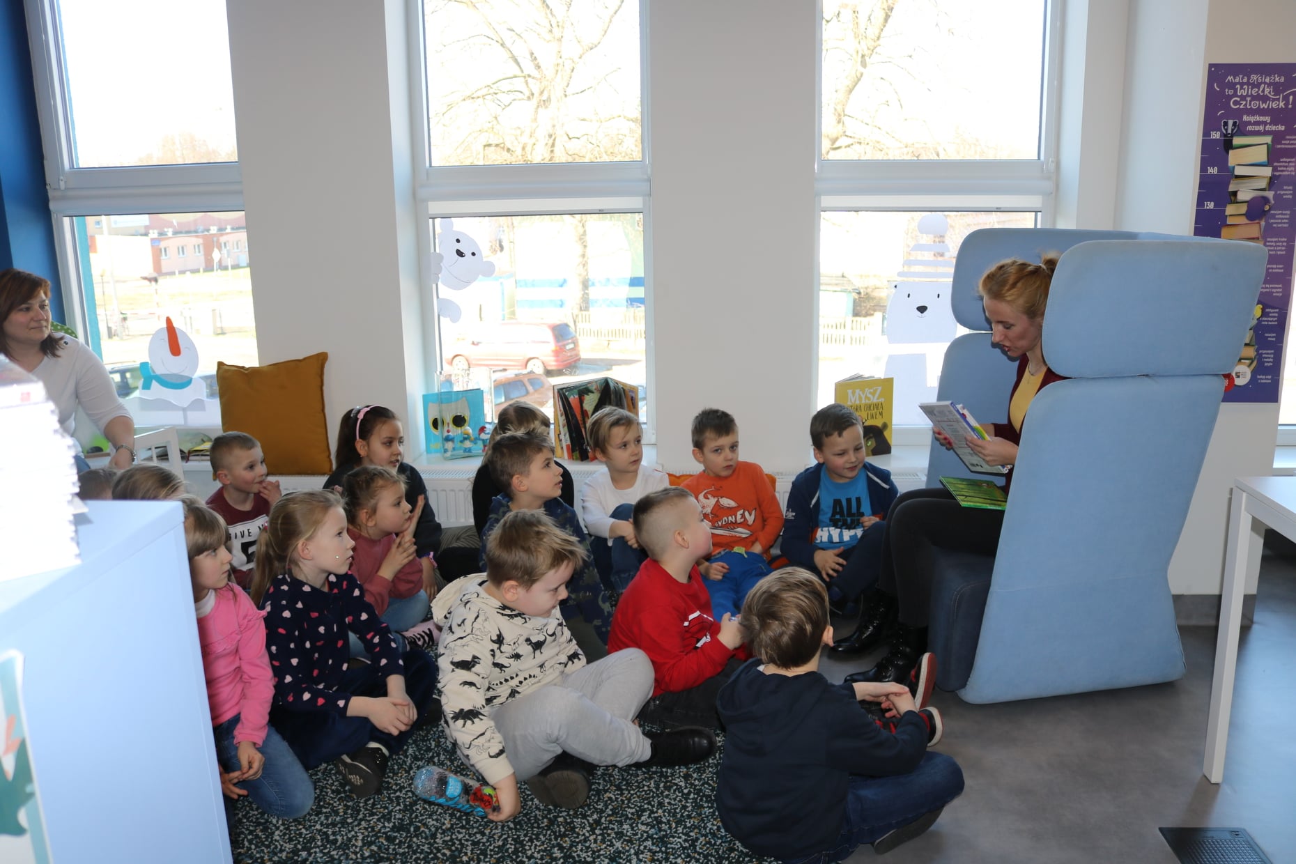 Dzieci siedzące na dywanie, słuchające bajke czytaną przez bibliotekarkę